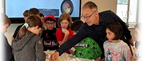 Eine Gruppe von Kindern und ein erwachsener Mann sind um einen Tisch versammelt und betrachten eine große Karte. Der Erwachsene erklärt etwas. Hinter ihnen befindet sich ein großer Bildschirm.