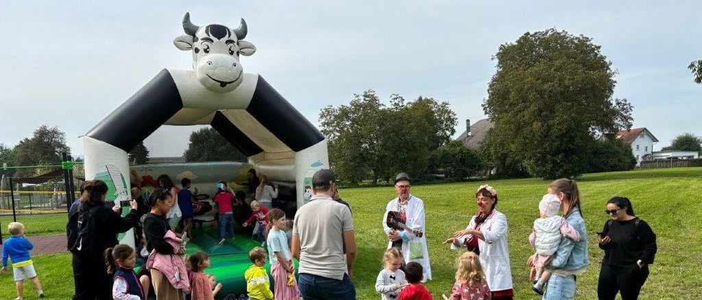 Eine Gruppe von Menschen, einschließlich Kindern, versammelt sich um eine aufblasbare Kuhstruktur auf einer Rasenfläche. Einige sind in Kostümen gekleidet und andere machen Fotos.