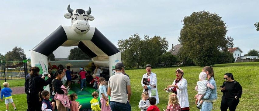 Eine Gruppe von Menschen, einschließlich Kindern, versammelt sich um eine aufblasbare Kuhstruktur auf einer Rasenfläche. Einige sind in Kostümen gekleidet und andere machen Fotos.