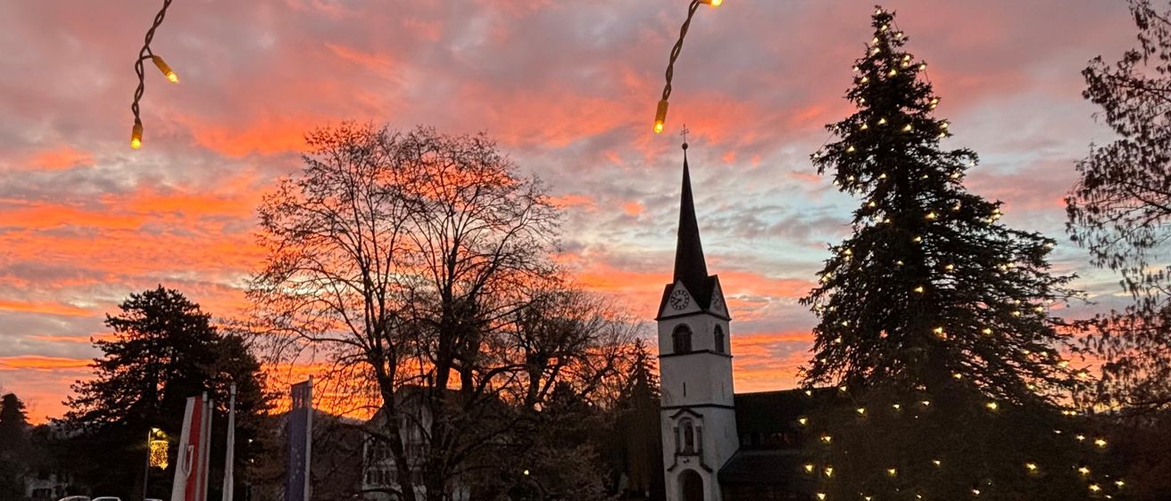 Ein Blick auf den Sonnenuntergang zeigt eine Kirche mit Turm und Spitze, umgeben von Bäumen und geschmückt mit Weihnachtslichtern. Eine dekorative Lichterkette hängt über dem Dach.