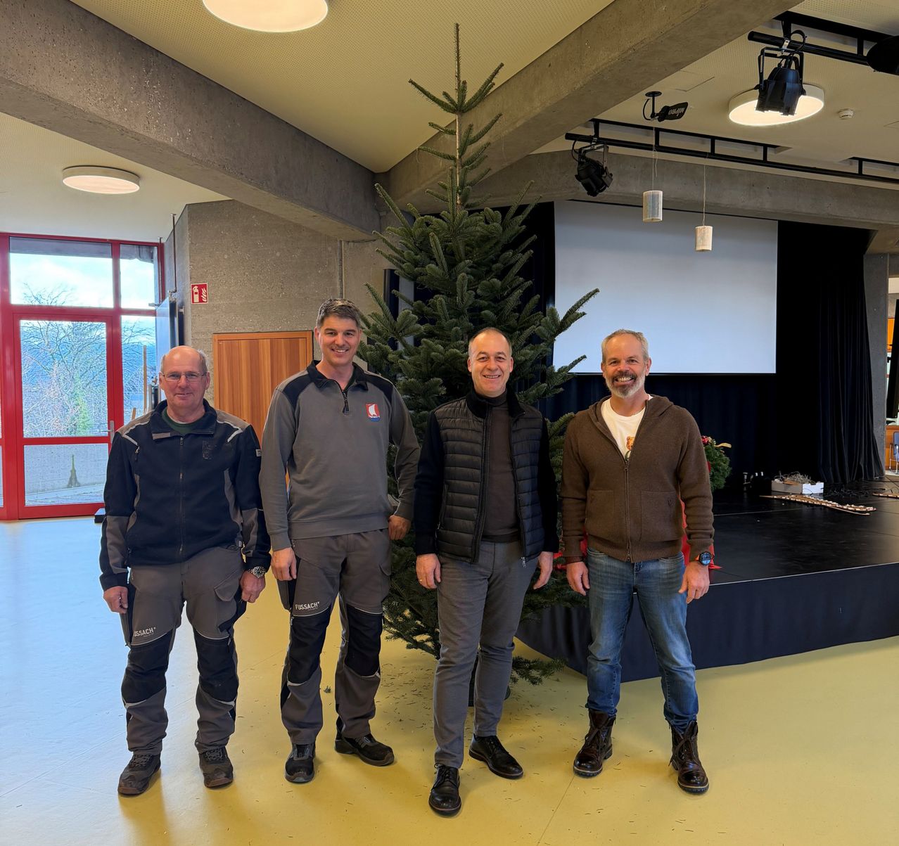 Vier Männer stehen vor einem Weihnachtsbaum in einem Raum mit Bühne, großem Fenster und Leinwand.