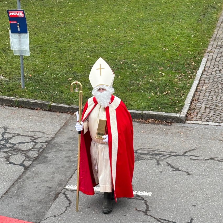 Eine Person, die als Bischof gekleidet ist, geht auf der Straße und hält einen goldenen Stab und ein Buch. Ein Schild mit dem Wort 'NEUE' ist links sichtbar.