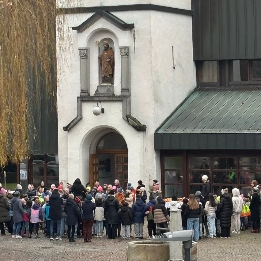 Eine Menschenmenge, darunter auch Kinder, steht vor einem Kirchengebäude und wartet in einer Schlange. Die Kirche hat eine Statue eines Heiligen und eine Lampe über dem Eingang.