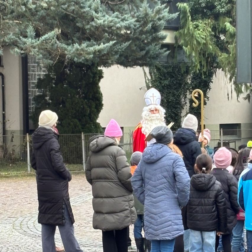 Eine Gruppe von Menschen, darunter Kinder, versammelt sich um einen als Weihnachtsmann verkleideten Mann auf einem Innenhof, mit Bäumen und einem Gebäude im Hintergrund.