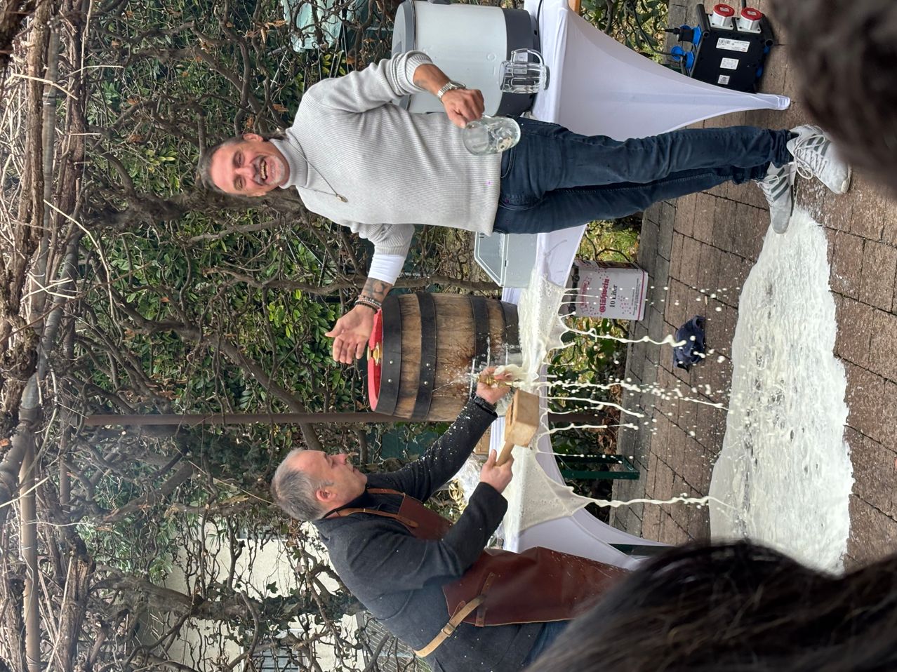 Zwei Männer gießen etwas in einen Fass, einer in Grau und einer in Braun. Die Flüssigkeit läuft über die Seiten.