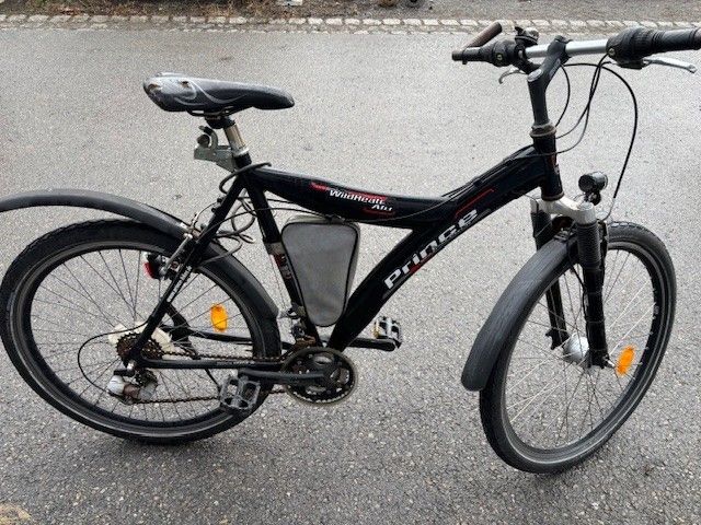 Ein schwarzes Fahrrad mit dem Wort Prince auf seinem Rahmen steht am Straßenrand. Das Fahrrad hat zwei Reflektoren, einen Flaschenhalter und eine Kette.