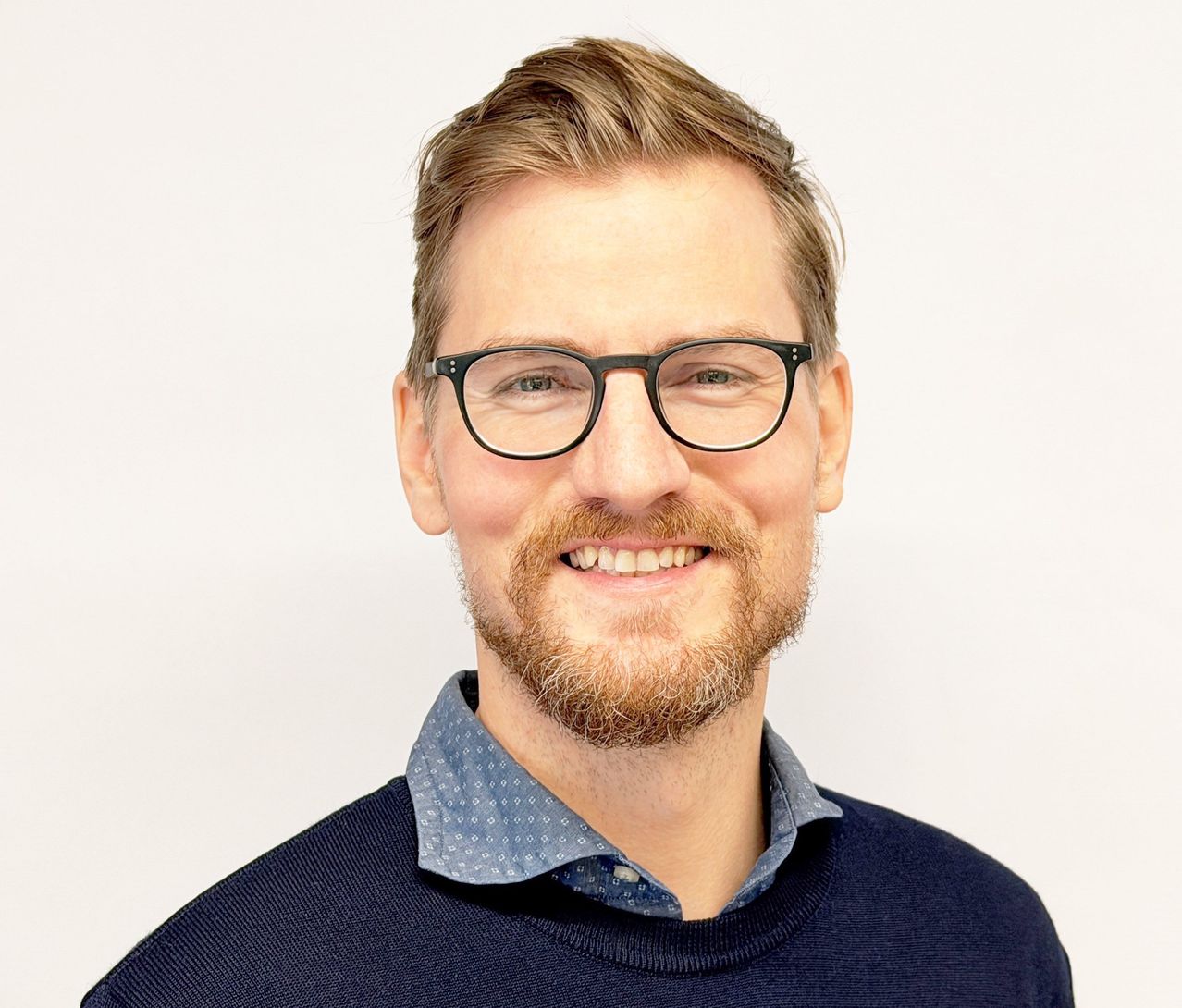 Ein Mann mit Bart und Brille lächelt und schaut in die Kamera. Er trägt einen marineblauen Pullover und ein hellblaues Hemd.