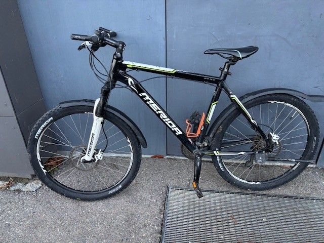 A black and white Merida mountain bike is parked on the side of a blue wall. The bike has two wheels and a seat.