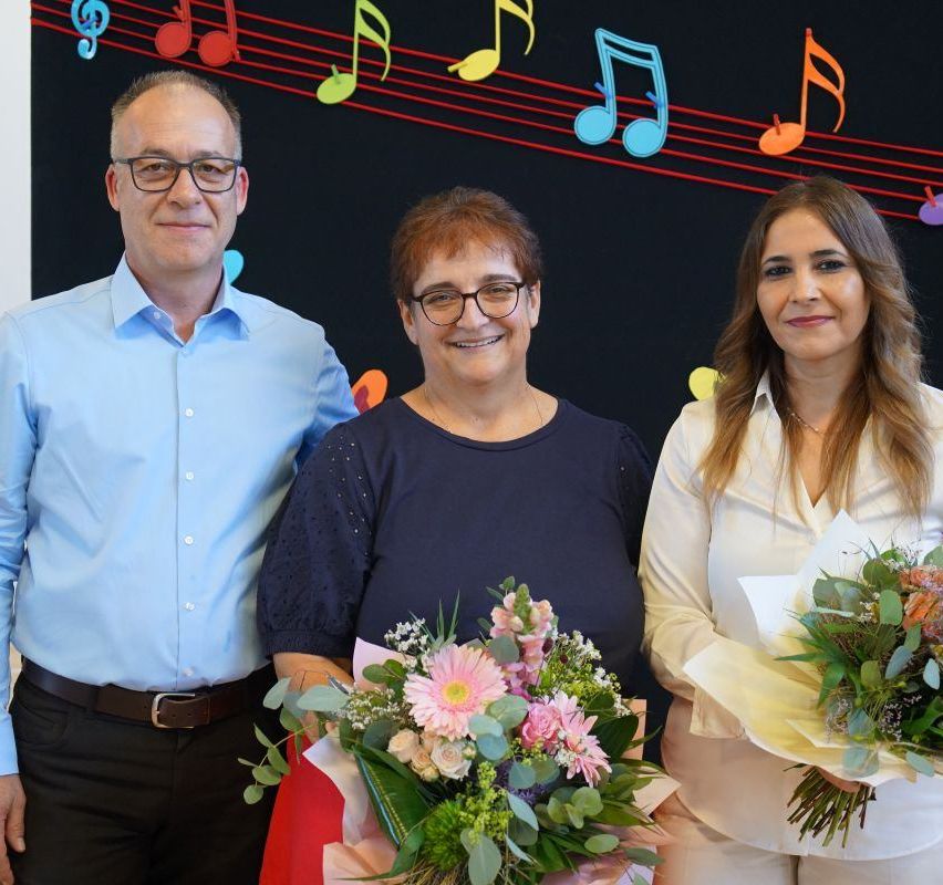 Three adults stand close together, smiling, in front of a wall with colorful musical notes. The man wears glasses and a blue shirt. The woman in the middle holds a large bouquet of flowers.