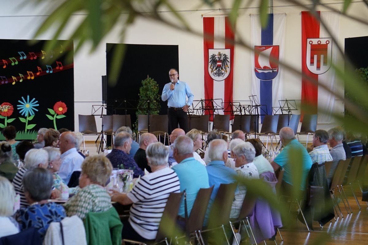 A man speaks into a microphone on stage in front of an audience. Behind him, banners are displayed, one featuring a coat of arms.