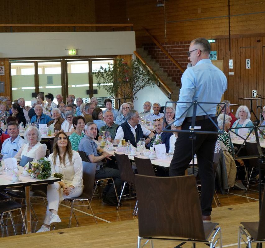 A large group of people are seated at tables in an indoor venue. A man stands in front of them, holding a microphone. The tables are set with flowers and glasses. Some people are wearing glasses. The room has wooden walls and a staircase.