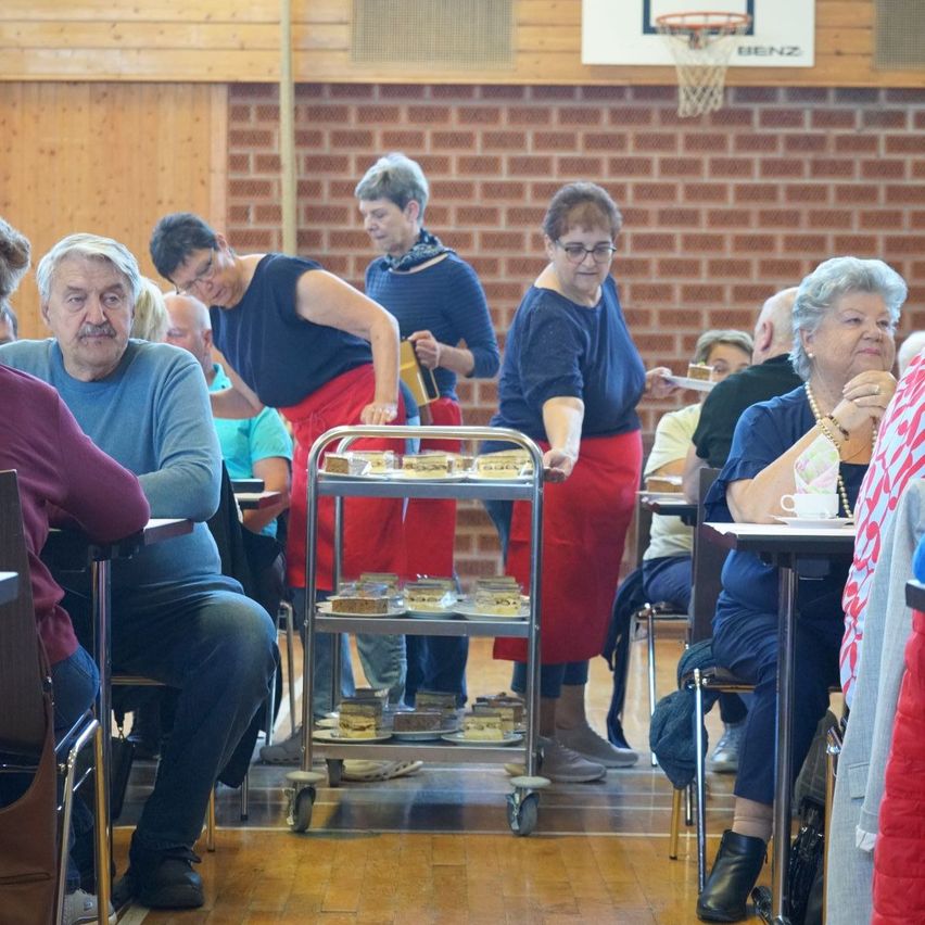 A group of elderly people sit at tables in a hall. Two women in aprons are serving food on a rolling cart. A basketball hoop is mounted on the brick wall.