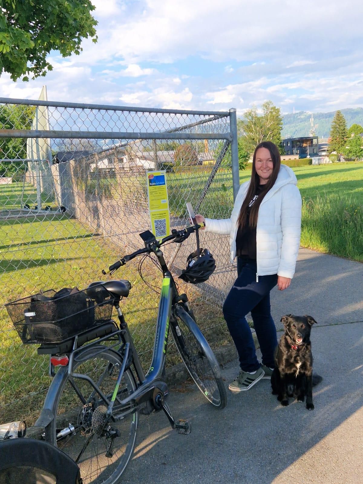 Eine Frau mit einer weißen Jacke steht neben einem Fahrrad, hält ein Schild mit einem QR-Code und steht neben einem Zaun mit einem sitzenden Hund. Dahinter befindet sich ein Grasbereich mit einem Gebäude und Bergen.