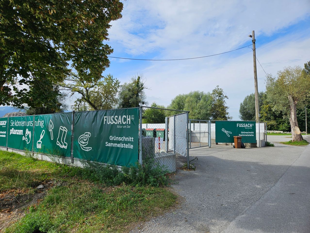Ein grüner Zaun mit Fussach Natur am See-Schildern steht in einem Park, mit einem offenen Tor und einem Mülleimer in der Nähe. Bäume und ein Versorgungsmast sind im Hintergrund.