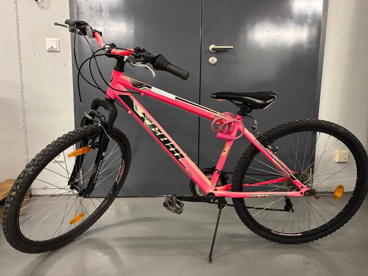 Ein rosafarbenes Fahrrad mit schwarzen Lenkern und Rädern steht vor einer grauen Tür. Das Fahrrad hat einen schwarzen Sattel und einen rosa Rahmen mit dem Wort 'HI-TEN' darauf.