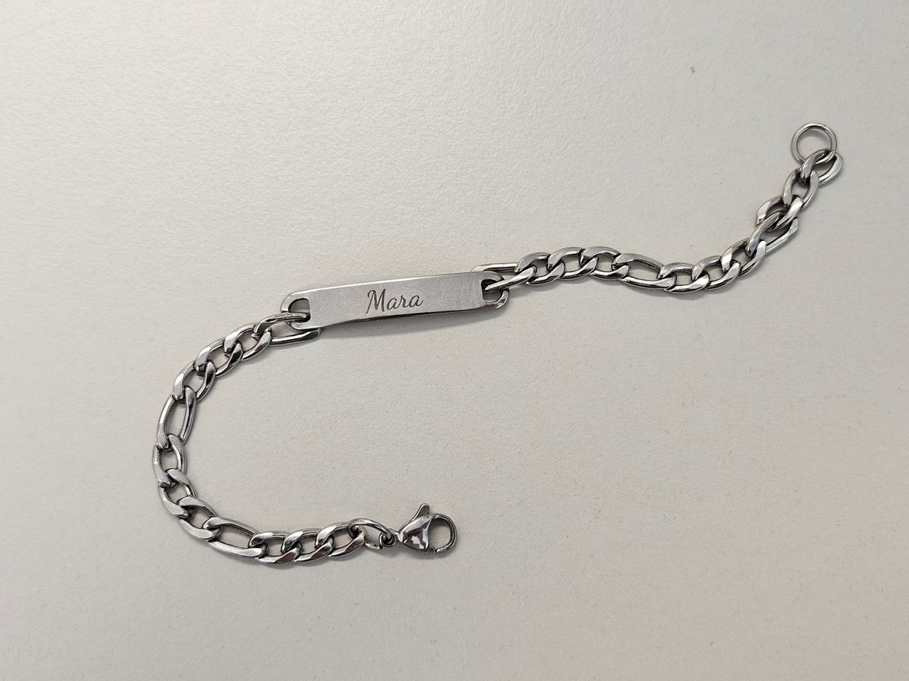 Eine silberne Armbandkette mit einem rechteckigen Balken, auf dem der Name 'Mara' eingraviert ist. Das Armband liegt auf einem hellen Hintergrund.