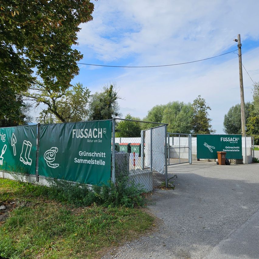 A green banner with the text 'FUSSAC' and 'Grünschnitt Sammelstelle' is displayed on a fence. The area is surrounded by trees and grass. There is a trash bin and a pole with wires. The sky is blue with some clouds.