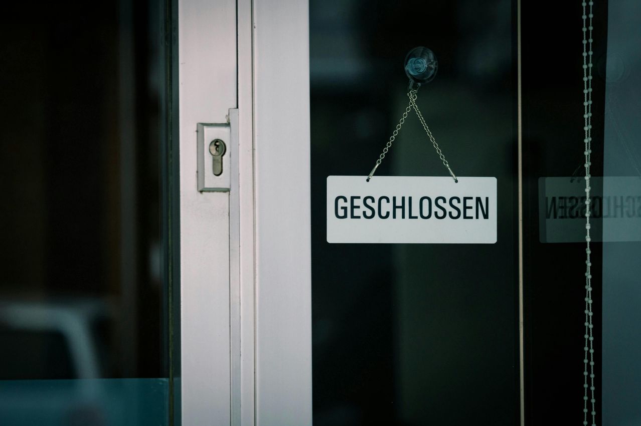 A white sign with black text reading 'Geschlossen' hangs on a glass door. The door is closed, and the lock is visible on the left side.