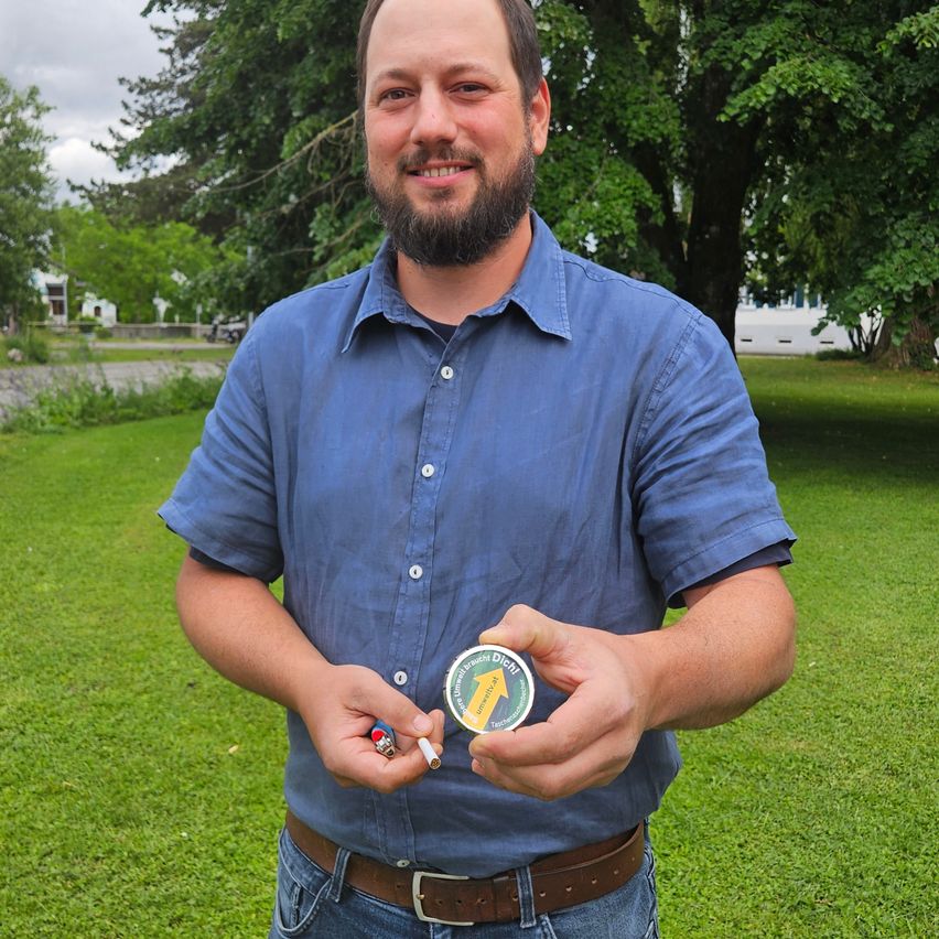 A man in a blue shirt stands outside, holding a small green container, a lighter, and a cigarette. The container reads 'Trusts Dich!' He smiles at the camera.