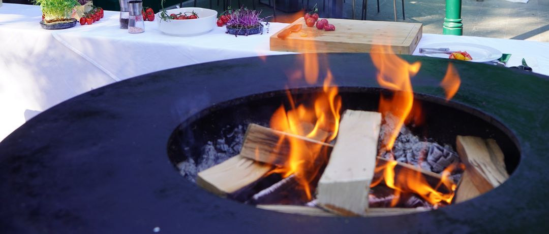 Eine Terrasse mit einem Feuerkorb und einem Tisch für eine Mahlzeit. Zwei Männer sitzen an einem Tisch, und eine Person steht an einer Bar. Der Bereich ist von grünen Säulen umgeben und hat eine weiße Tischdecke.
