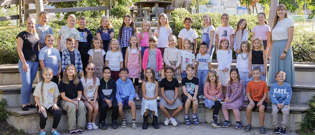 Eine Gruppe von Kindern und Jugendlichen posiert für ein Foto in einer Außenumgebung, umgeben von Grün und einer Holzstruktur.