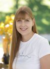 Eine Frau mit langen blonden Haaren lächelt für ein Foto. Sie trägt ein weißes T-Shirt mit dem Wort 'laie' darauf. Im Hintergrund ist eine verschwommene Pflanze mit gelben Blättern zu sehen.