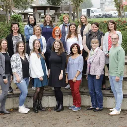 Eine Gruppe von Erwachsenen, sowohl Männer als auch Frauen, posiert für ein Foto im Freien. Sie stehen vor einer Betonmauer mit Pflanzen und Grünpflanzen dahinter.
