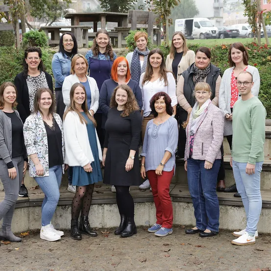 Eine Gruppe von Erwachsenen, sowohl Männer als auch Frauen, posiert für ein Foto im Freien. Sie stehen vor einer Betonmauer mit Pflanzen und Grünpflanzen dahinter.