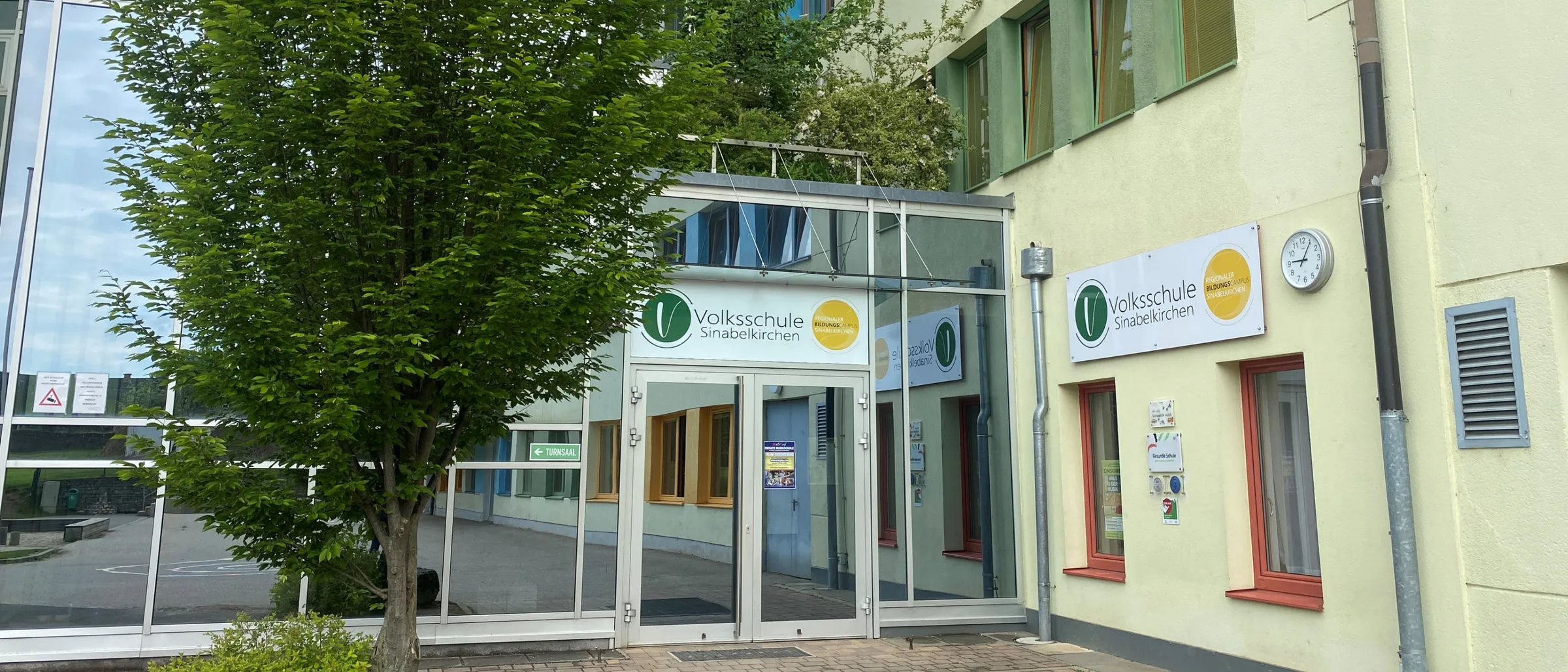 Der Eingang zu einem Gebäude verfügt über eine Glasüberdachung, einen Baum und eine Uhr. Das Gebäude hat gelbe und grüne Fenster und ein Schild mit der Aufschrift Volksschule Sinabelkirchen.