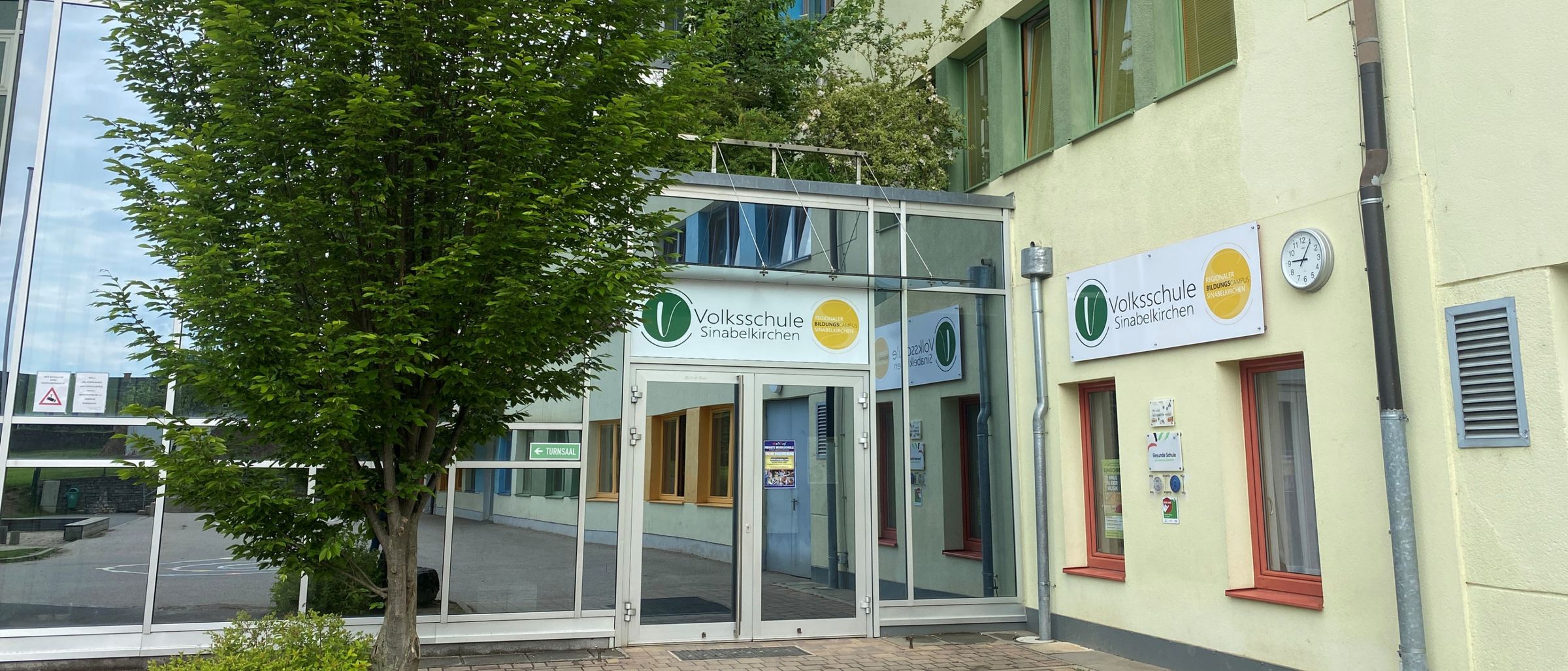 Der Eingang zu einem Gebäude verfügt über eine Glasüberdachung, einen Baum und eine Uhr. Das Gebäude hat gelbe und grüne Fenster und ein Schild mit der Aufschrift Volksschule Sinabelkirchen.