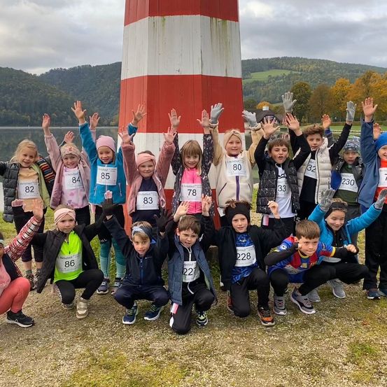 Eine Gruppe von Kindern posiert für ein Foto mit Nummern auf ihren Shirts, neben einem Leuchtturm, mit einem See und Hügeln im Hintergrund.