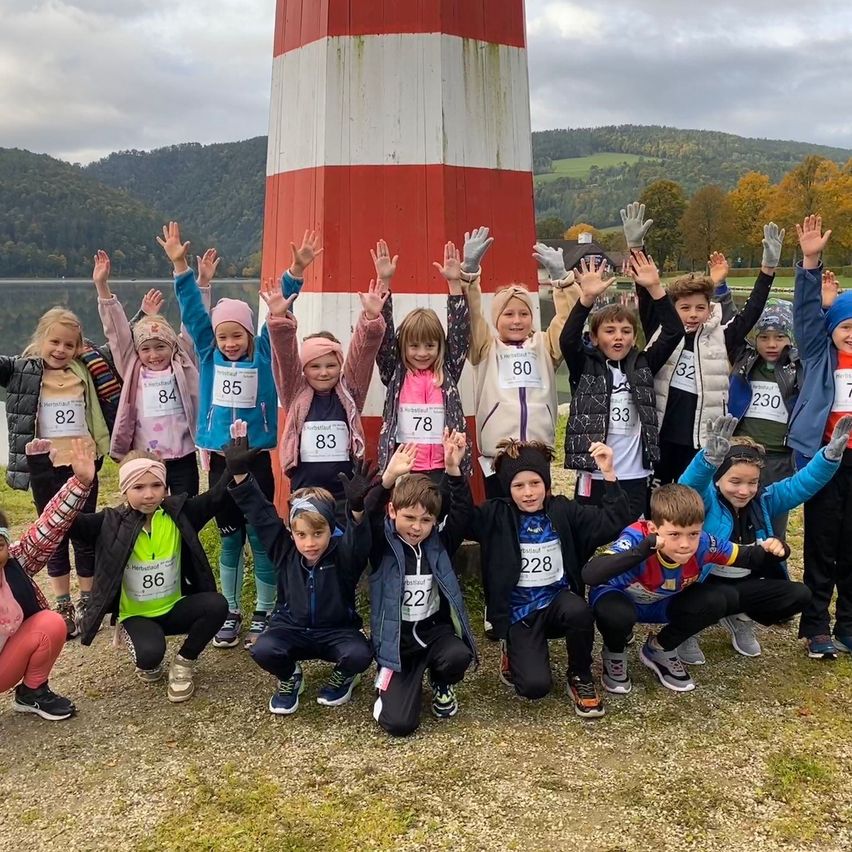 Eine Gruppe von Kindern posiert für ein Foto mit Nummern auf ihren Shirts, neben einem Leuchtturm, mit einem See und Hügeln im Hintergrund.