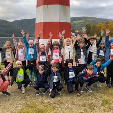 Eine Gruppe von Kindern posiert für ein Foto mit Nummern auf ihren Shirts, neben einem Leuchtturm, mit einem See und Hügeln im Hintergrund.