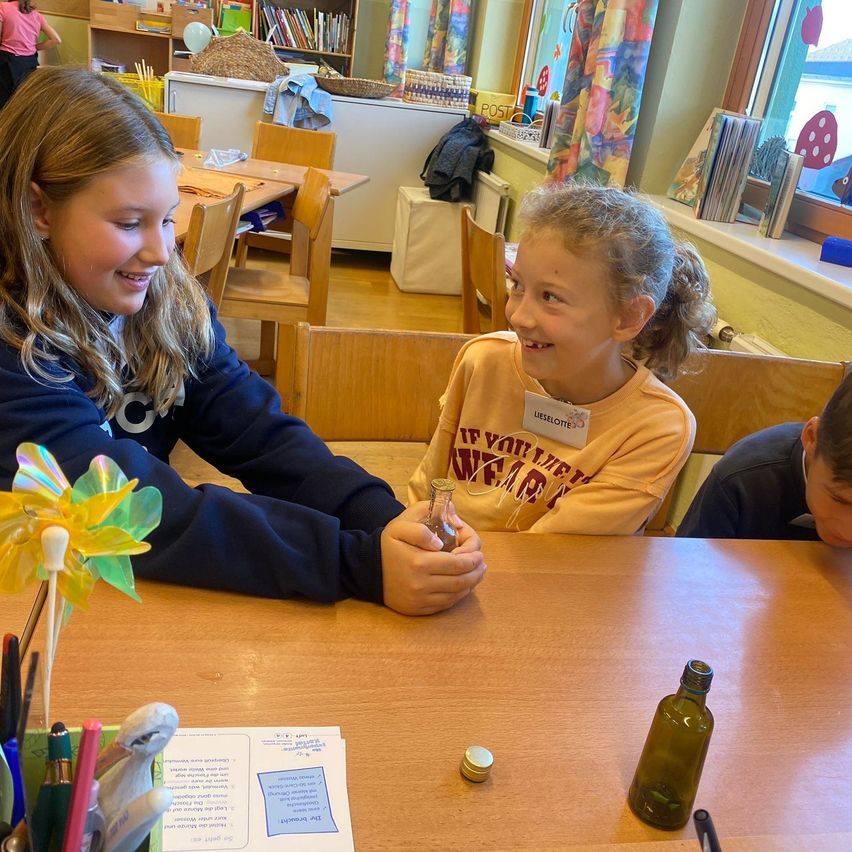 Zwei junge Mädchen lächeln und halten eine kleine Flasche, sie sitzen an einem Tisch mit einem Windrad, einem Stift und einem kleinen Glas. Ein Junge sitzt hinter ihnen.
