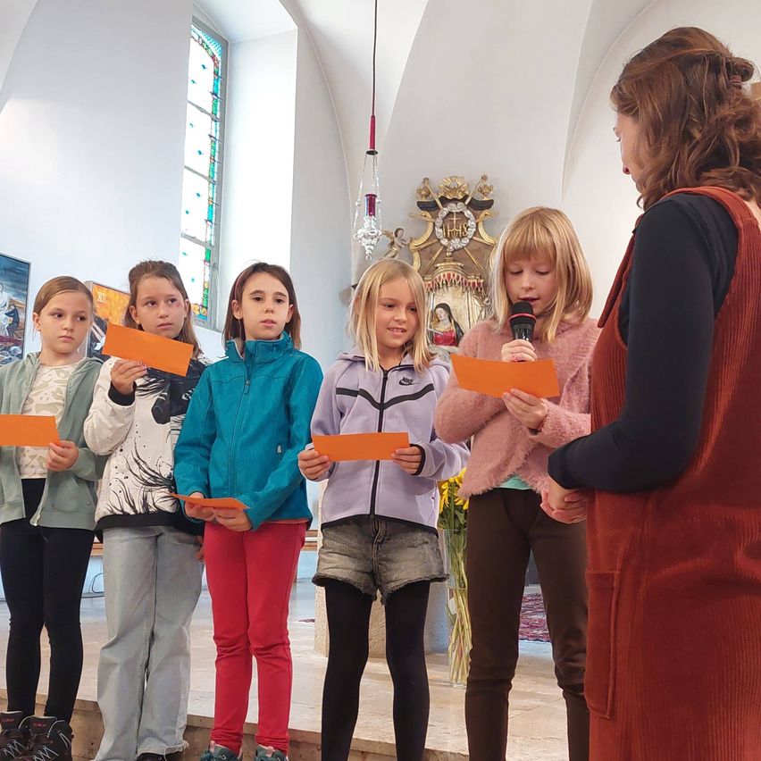 Eine Gruppe junger Mädchen hält orangefarbene Papiere in einer Kirche. Eine Frau hält ein Mikrofon, und ein Blumenvase steht in der Nähe.