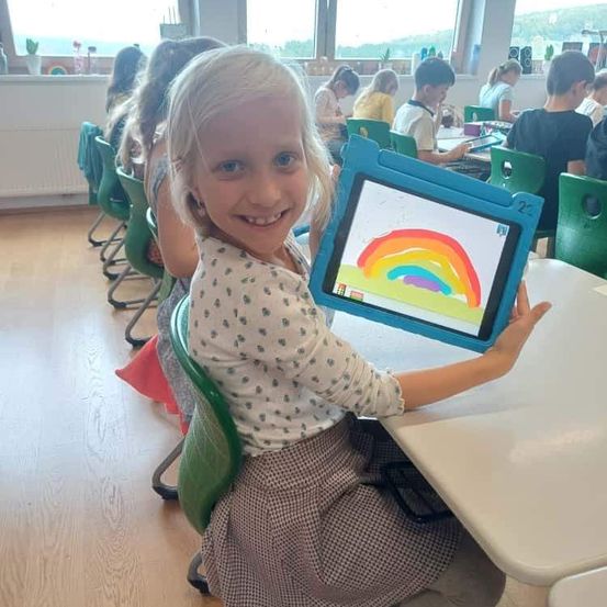 Ein kleines Mädchen sitzt in einem Klassenzimmer und hält ein Tablet mit einer Regenbogenzeichnung. Andere Kinder sitzen um sie herum, und ein weißer Tisch steht vor ihnen.