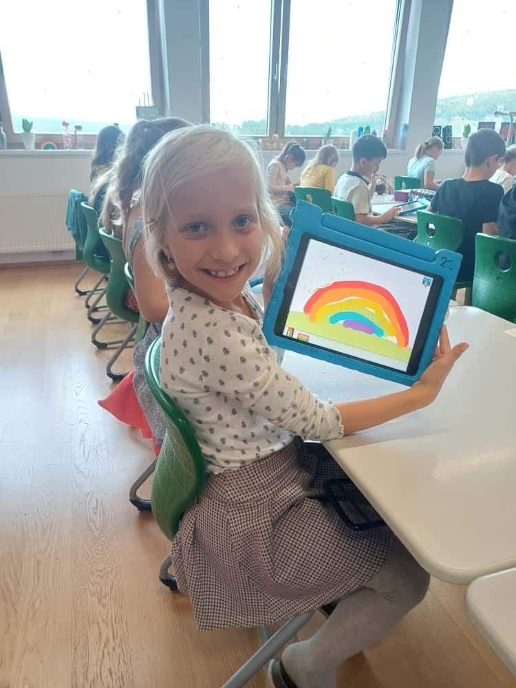 Ein kleines Mädchen sitzt in einem Klassenzimmer und hält ein Tablet mit einer Regenbogenzeichnung. Andere Kinder sitzen um sie herum, und ein weißer Tisch steht vor ihnen.