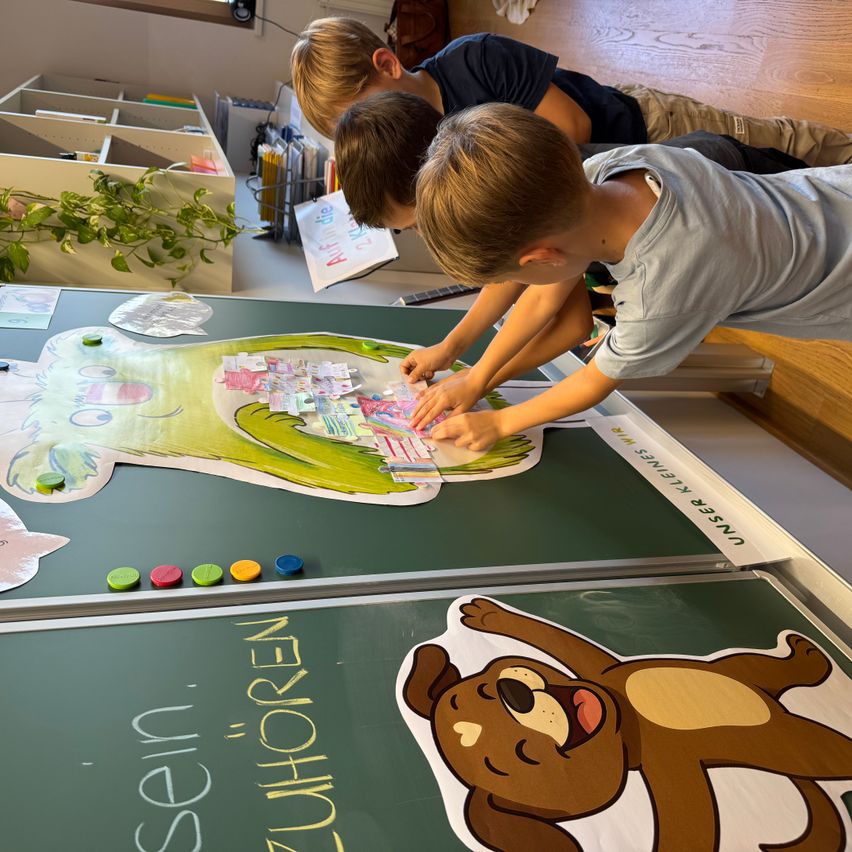 Drei Kinder spielen mit einem großen grünen Froschpuzzle auf einer Tafel. Zwei Kinder sitzen, während eines steht und am Puzzle arbeitet.