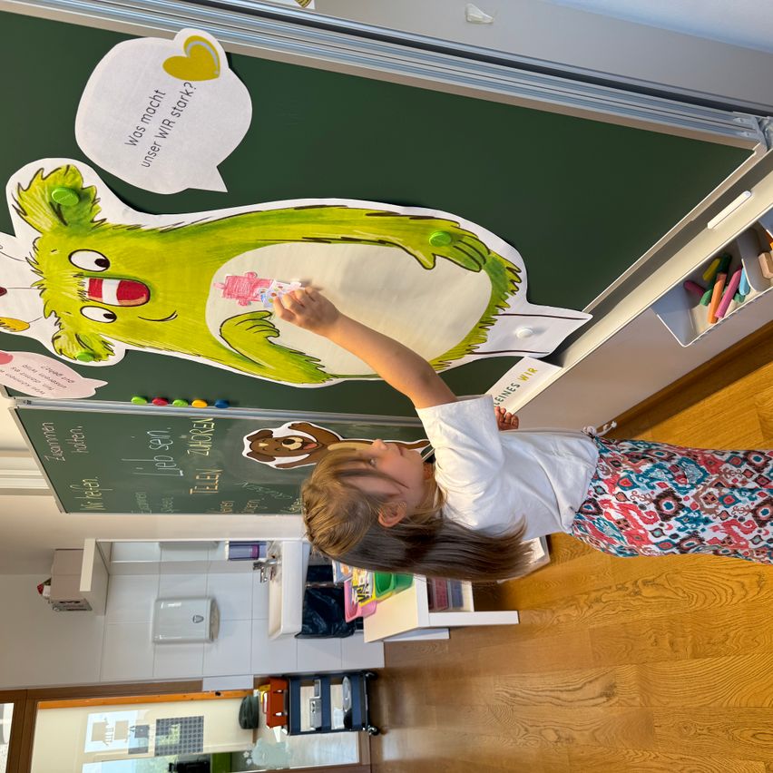 Ein kleines Kind heftet eine grüne Monster-Papiervorlage an eine Tafel mit deutschem Text, umgeben von einer Klassenraumumgebung.