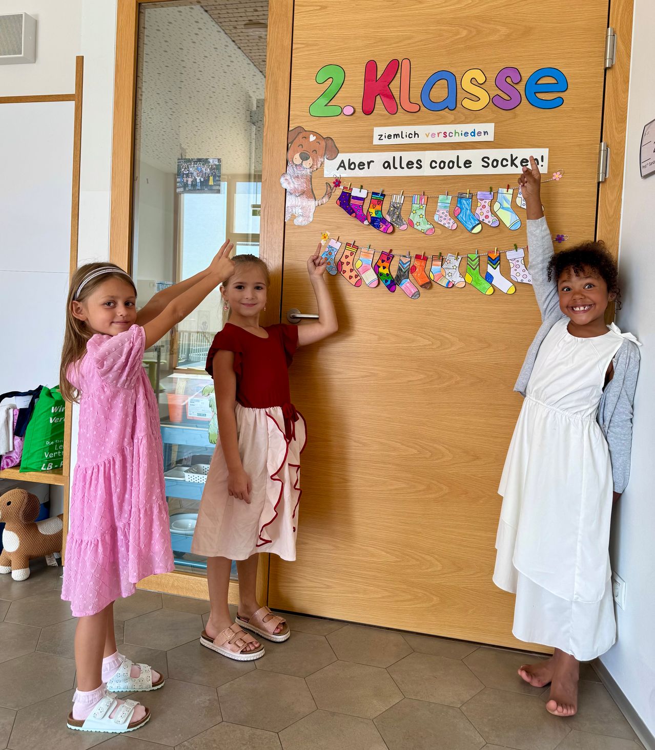Drei junge Mädchen stehen an einer Tür mit einem Schild, auf dem 2. Klasse steht. Ein Mädchen berührt die bunten Socken, die an einer Wäscheleine über der Tür hängen.