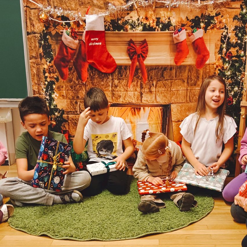 Eine Gruppe von Kindern sitzt um einen Kamin herum, jeder hält ein Geschenk. Im Hintergrund hängen Weihnachtsdekorationen und Strümpfe.