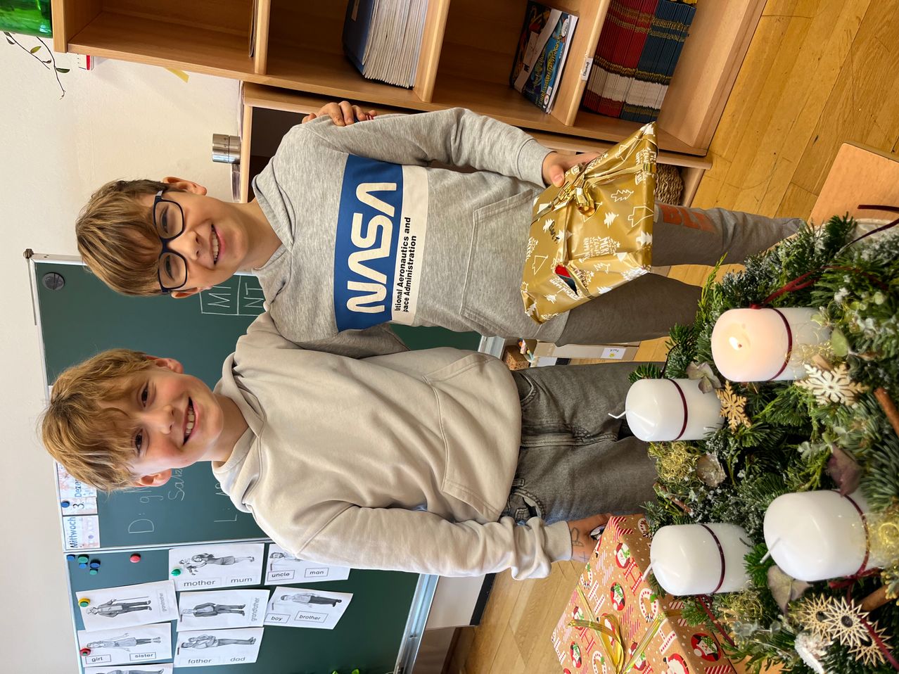 Zwei Jungen lächeln in einem Klassenzimmer mit einem Weihnachtsbaum, Geschenken und einer Tafel mit Familienwörtern. Ein Junge trägt ein NASA-Sweatshirt und hält ein Geschenk.