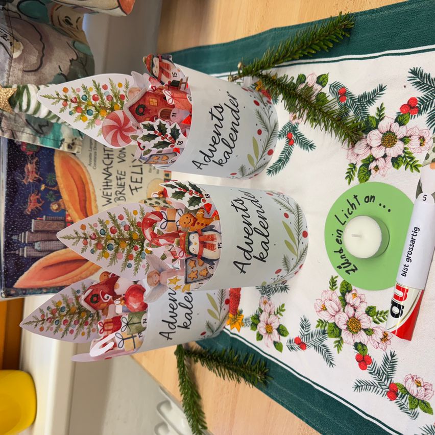 Eine Tischdekoration mit Weihnachtsdekoration, einschließlich Tassen mit Weihnachtsszenen, einer Kerze und einem Kranz auf einer Tischdecke. Es gibt auch Tannenzweige und Blumen.