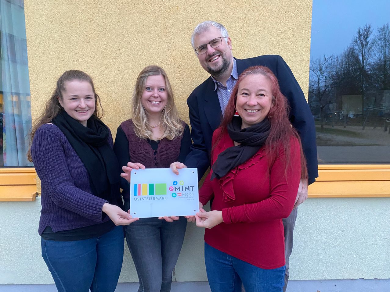 Vier Menschen stehen lächelnd vor einem gelben Gebäude und halten ein Schild mit der Aufschrift 'MINT' und 'OSTSTEIERMARK'. Zwei Frauen tragen Schals und der Mann in der Mitte trägt eine Brille.