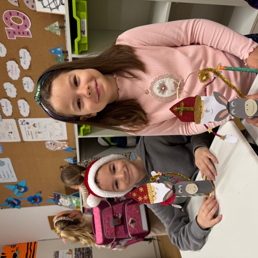 Zwei Kinder sind in einem Klassenzimmer, eines hält eine Papierbastelarbeit eines Esels und lächelt. Ein anderes Kind, das einen Weihnachtsmannhut trägt, sitzt und hält eine Papierbastelarbeit eines Heiligen und eines Esels. Hinter ihnen zeigt eine Pinnwand verschiedene Papiere und Dekorationen.