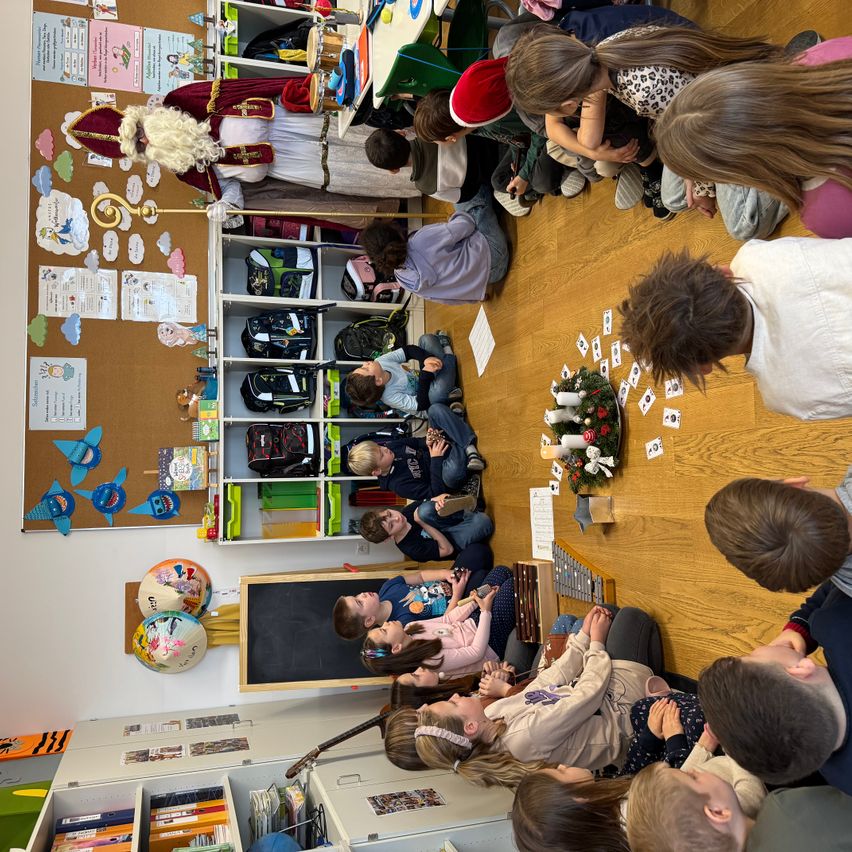 Ein Klassenzimmer mit Kindern, die um einen Weihnachtskranz herum auf dem Boden sitzen, einer als Weihnachtsmann verkleidet. Sie scheinen an einer festlichen Aktivität teilzunehmen.