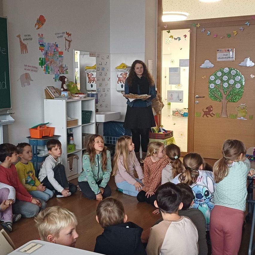 Eine Lehrerin steht vor einem Klassenzimmer voller Kinder und hält eine Tablett. Das Klassenzimmer hat eine Tafel und einen Schreibtisch mit Regalen, mit verschiedenen Dekorationen und Spielzeugen drumherum.