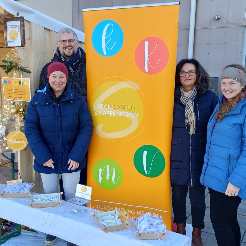 Vier Personen stehen vor einem Tisch mit Essen. Dahinter befindet sich ein Banner mit bunten Buchstaben und den Worten 'Regio Campus Sinabelkirchen'.