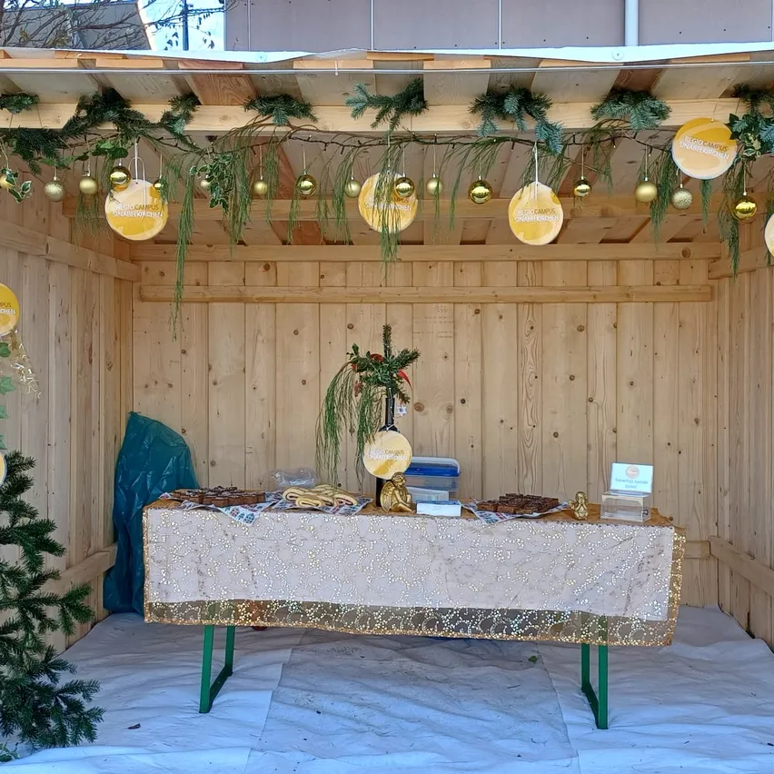 Eine festliche Weihnachtseinrichtung mit Holzwänden und einem Tisch, der mit einer goldenen Tischdecke bedeckt ist. Verschiedene Gegenstände sind auf dem Tisch und ein Weihnachtsbaum steht links.