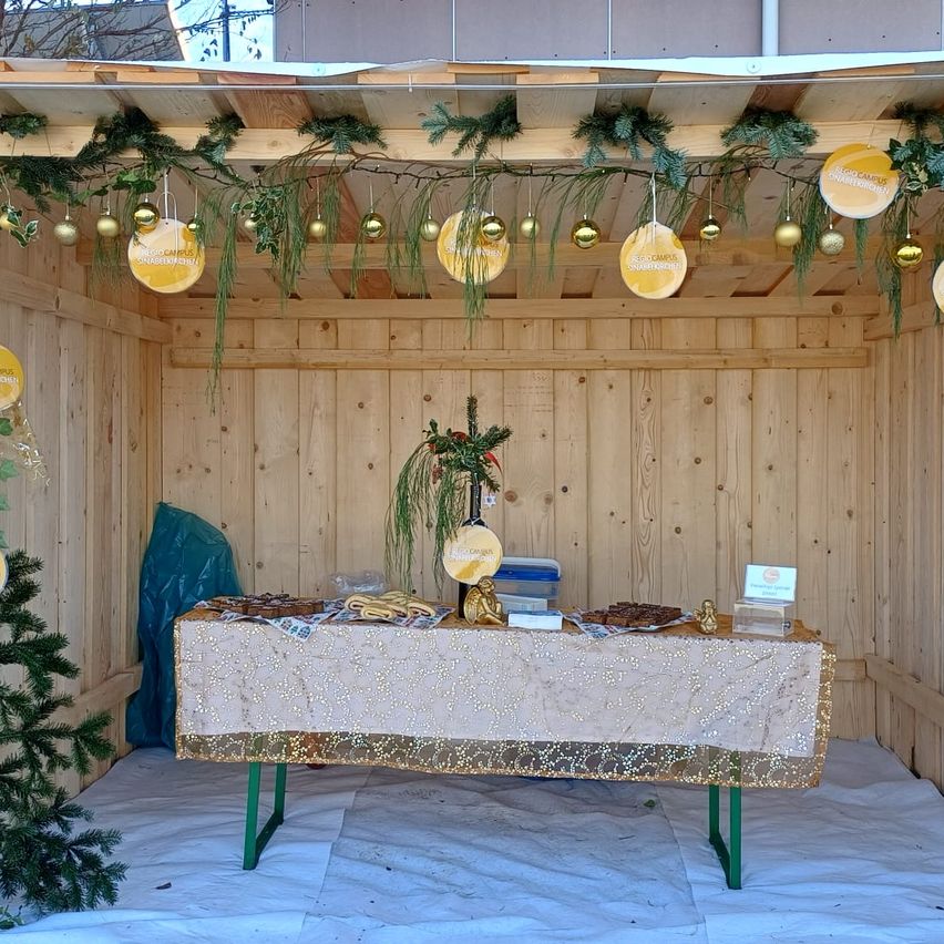 Eine festliche Weihnachtseinrichtung mit Holzwänden und einem Tisch, der mit einer goldenen Tischdecke bedeckt ist. Verschiedene Gegenstände sind auf dem Tisch und ein Weihnachtsbaum steht links.